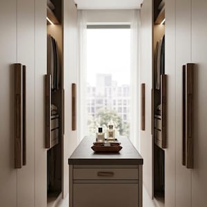 May include: A walk-in wardrobe featuring floor-to-ceiling beige cabinets with vertical dark wood handles, a central island with a dark countertop holding two glass perfume bottles, and a beige upholstered ottoman positioned in the foreground.