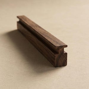 May include: A long, rectangular piece of dark brown walnut wood with a visible natural grain pattern. The object features a distinct horizontal groove running along its top edge, creating a T-shaped profile when viewed from the side end.