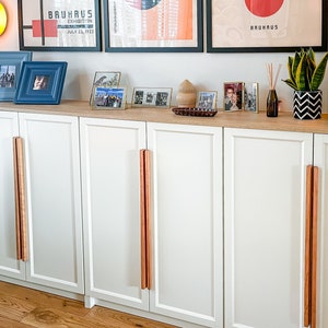 May include: White cabinet with four doors and wooden pulls. The cabinet is decorated with framed photos and a potted plant.