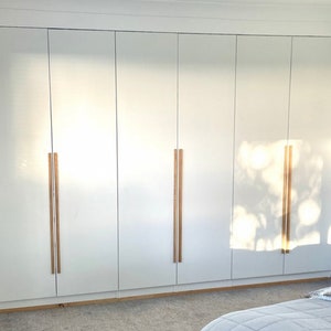 May include: A large, floor-to-ceiling white wardrobe unit featuring six flat-panel doors. Each pair of doors is fitted with long, vertical, light-toned wooden bar handles. The smooth white finish reflects light, creating bright highlights on the surface.
