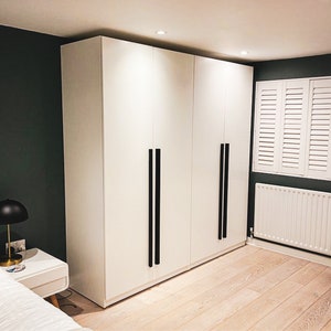 May include: A large, floor-to-ceiling white wardrobe featuring four smooth, flat-panel doors. Each pair of doors is fitted with long, vertical, matte black metal handles that extend down the centre, providing a stark contrast to the white finish.