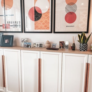 May include: A white cabinet with three doors and wooden pulls. The cabinet is decorated with three framed posters with geometric designs and the text "BAUHAUS" and "FÜHREND IN DER HERRENMODE". There are also several framed photos on top of the cabinet.