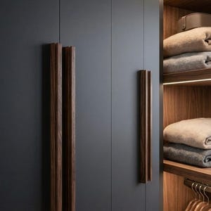 May include: A dark gray wardrobe with vertical wooden handles and an open shelving section. The interior features warm lighting, wooden shelves holding folded beige and gray textiles, and a rack with wooden hangers for clothing storage.