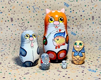 May include: Set of five hand-painted wooden nesting dolls, each depicting a cat with different colors and designs. The largest doll is orange with a fish design, the second largest is gray, the third is gray and white with a red and white polka dot pattern, the fourth is gray and white, and the smallest is gray and white with a red and white polka dot pattern.