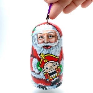 May include: A hand-painted Christmas ornament shaped like Santa Claus, with a red coat and white beard. The ornament features a painted image of a nutcracker. A purple ribbon is attached for hanging.