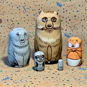 Può includere: Un set di cinque bambole russe in legno dipinte a mano, o bambole matrioska, che raffigurano un orso, una volpe, un procione, uno scoiattolo e un topo. Le bambole sono di dimensioni diverse e sono dipinte con colori realistici.