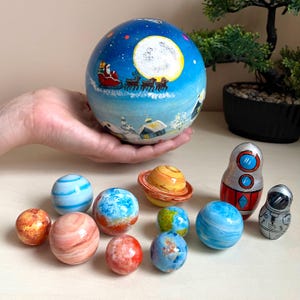 May include: A collection of hand-painted wooden nesting dolls and planet-themed spheres. The large egg-shaped doll depicts a winter scene with Santa's sleigh. The smaller spheres represent planets, and there are also astronaut and rocket ship dolls.