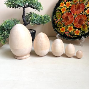 May include: Five wooden nesting eggs in various sizes, all painted in a natural wood color. The eggs are arranged in a row from largest to smallest.