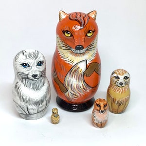 Puede incluir: Un conjunto de cinco muñecas rusas de anidación, o muñecas matrioska, con diseños de animales pintados. La muñeca más grande es un zorro rojo, la siguiente es un zorro ártico blanco, seguida de una suricata marrón, un zorro rojo más pequeño y la muñeca más pequeña es un pájaro marrón y blanco.