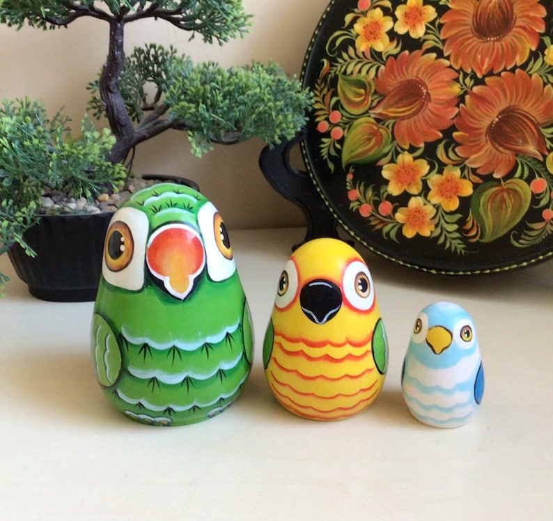 Peut inclure: Trois poup&eacute;es gigognes en bois peintes &agrave; la main en forme d'oiseaux. La plus grande poup&eacute;e est verte avec un bec rouge et des yeux jaunes. La poup&eacute;e du milieu est jaune avec un bec noir et des yeux rouges. La plus petite poup&eacute;e est bleue avec un bec jaune et des yeux blancs.