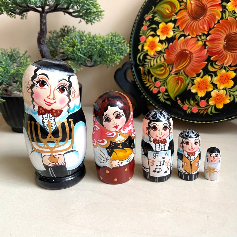 以下が含まれることがあります： 手描きの入れ子人形6体のセット。最大の人形は、金色のメノラーを持つ人物を描いています。人形は詳細な顔の表情をしており、伝統的な衣装を着ています。背景には装飾的なトレイがあります。