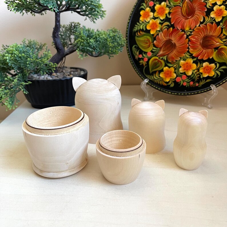 May include: Set of five unfinished wooden nesting dolls, each with a cat-shaped head and a smooth, rounded body. The dolls are arranged in decreasing size, with the largest doll in the back and the smallest doll in the front.