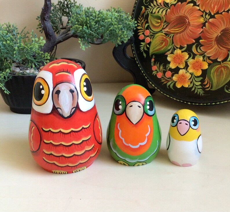 Peut inclure: Trois poup&eacute;es russes en bois peintes &agrave; la main en forme d'oiseaux. La plus grande poup&eacute;e est rouge avec des accents orange et jaunes, la poup&eacute;e du milieu est verte avec des accents orange et jaunes, et la plus petite poup&eacute;e est jaune avec des accents blancs.