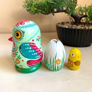 May include: A set of three hand-painted wooden nesting dolls. The largest doll is shaped like a green bird with pink, blue, and yellow details. The middle doll is shaped like a white egg with green grass and flowers. The smallest doll is shaped like a yellow chick with red, white, and blue details.