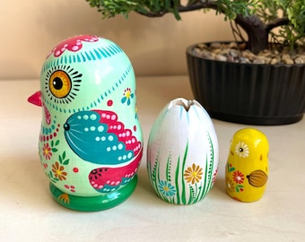 Könnte beinhalten: Ein Set aus drei handbemalten, ineinander verschachtelten Holzpuppen. Die größte Puppe ist in Form eines grünen Vogels mit rosa, blauen und gelben Details gestaltet. Die mittlere Puppe ist in Form eines weißen Eies mit grünem Gras und Blumen gestaltet. Die kleinste Puppe ist in Form eines gelben Küken mit roten, weißen und blauen Details gestaltet.