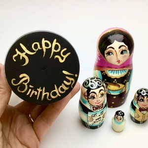 以下が含まれることがあります： 「Happy Birthday!」と金色の文字が入った黒い丸いオブジェクトが、ロシアのマトリョーシカ人形のセットの隣に持たれています。人形は、さまざまな色とデザインで、詳細な顔と服で塗装されています。