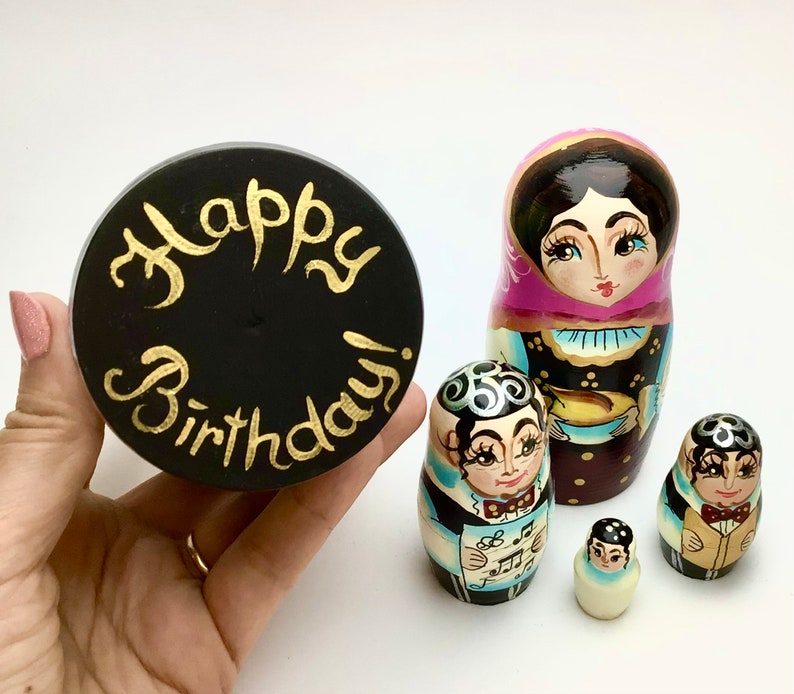 以下が含まれることがあります： 「Happy Birthday!」と金色の文字が入った黒い丸いオブジェクトが、ロシアのマトリョーシカ人形のセットの隣に持たれています。人形は、さまざまな色とデザインで、詳細な顔と服で塗装されています。