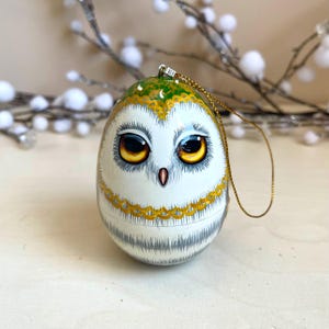 Peut inclure: Ornement en forme d'œuf peint à la main représentant un hibou. Il est blanc avec des détails jaunes et verts. Le hibou a de grands yeux avec des iris jaunes et une ficelle dorée. Le design est détaillé et texturé.