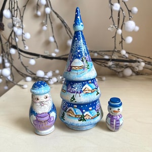 Könnte beinhalten: Ein Set aus drei handbemalten Weihnachtsornamenten aus Holz. Die Ornamente umfassen einen blau-weißen Weihnachtsbaum, eine Weihnachtsmannfigur und einen Schneemann. Baum und Weihnachtsmann sind mit einer Winterdorf-Szene bemalt.
