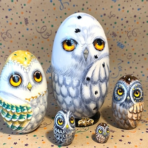 Peut inclure: Ensemble de cinq poupées russes en bois peintes à la main, chacune représentant un hibou différent avec des yeux jaunes. La plus grande poupée est blanche avec des marques grises, la suivante est jaune avec des marques vertes, la suivante est grise avec des marques brunes, et les deux plus petites sont grises avec des marques bleues.
