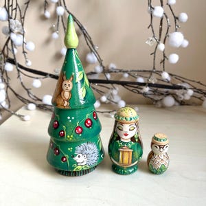 May include: A set of three hand-painted wooden figurines: a green Christmas tree with a squirrel and berries, a woman holding a lantern, and an owl. The figurines are painted in bright colors and have a folk art style.