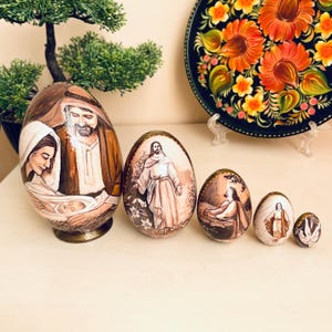 Peut inclure: Un ensemble de six œufs de Pâques décoratifs peints à la main. Les œufs présentent des scènes religieuses, notamment des représentations de Jésus, Marie et Joseph. Les œufs varient en taille et sont exposés sur une surface claire. Le plus grand œuf a une base en bronze.