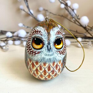 Könnte beinhalten: Handbemalter Eulen-Ornament. Die Eule hat braune, weiße und orangefarbene Federn mit großen Augen. Ein goldener Faden ist zum Aufhängen befestigt. Das Ornament ist ein dekorativer Gegenstand. Die Maße sind in Zentimetern.