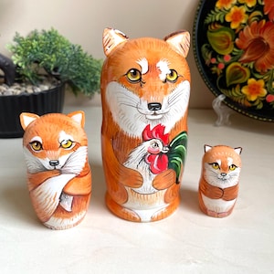May include: A set of three Russian nesting dolls, or matryoshka dolls, featuring a fox design. The largest doll is holding a red rooster. The dolls are painted in shades of orange, white, and brown.