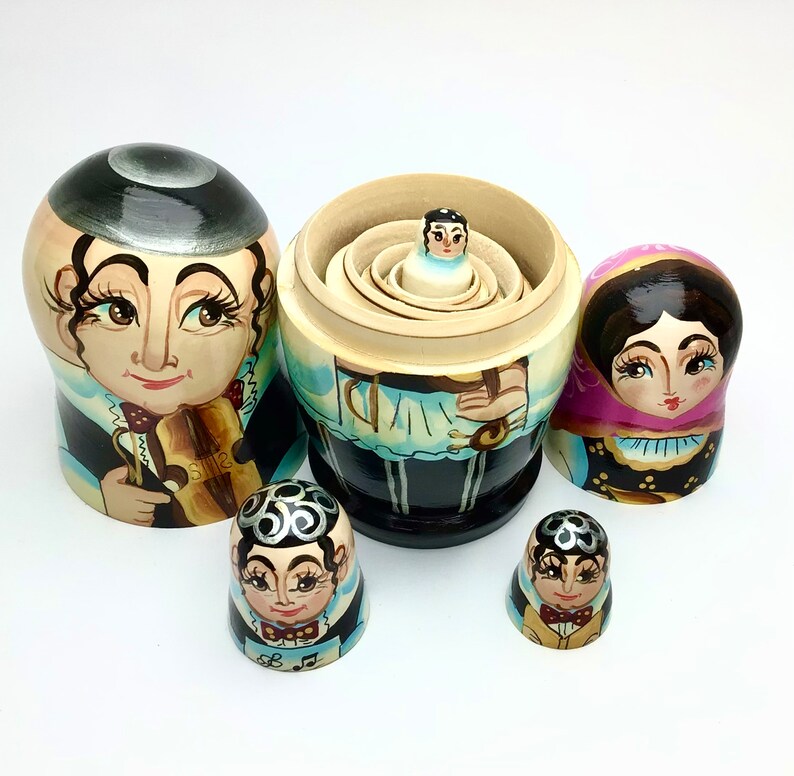 以下が含まれることがあります： 音楽をテーマにした、6体のロシアのマトリョーシカ人形のセット。人形は、バイオリニスト、ピンクのショールを着た女性、その他の人物が描かれた、詳細な顔と衣装で塗装されています。最大の人形は開いており、中には小さな人形が入っています。最大の高さは約15cmです。
