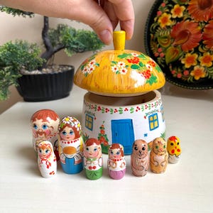 Könnte beinhalten: Ein Satz aus Holz-Nistpuppen, auch Matrjoschka-Puppen genannt, die im traditionellen russischen Stil bemalt sind. Die Puppen sind in einem Kreis um ein Holzhaus mit Strohdach angeordnet. Das Haus ist mit einer blauen Tür und Fenstern bemalt. Die Puppen sind alle unterschiedlich groß und haben verschiedene Designs, darunter eine Frau in einem blauen Kleid, ein Mann mit einem Schnurrbart, ein Mädchen mit einer Blumenkrone und eine Katze.