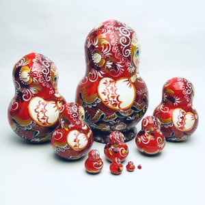 Puede incluir: Un conjunto de ocho mu&ntilde;ecas rusas tradicionales de anidaci&oacute;n, o mu&ntilde;ecas matrioska, pintadas con un patr&oacute;n floral rojo, blanco y dorado. La mu&ntilde;eca m&aacute;s grande mide aproximadamente 25 cm de alto y la m&aacute;s peque&ntilde;a aproximadamente 2,5 cm.