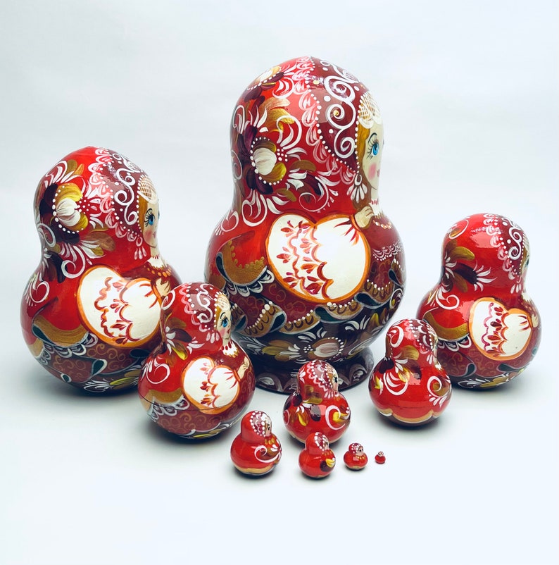 Puede incluir: Un conjunto de ocho mu&ntilde;ecas rusas tradicionales de anidaci&oacute;n, o mu&ntilde;ecas matrioska, pintadas con un patr&oacute;n floral rojo, blanco y dorado. La mu&ntilde;eca m&aacute;s grande mide aproximadamente 25 cm de alto y la m&aacute;s peque&ntilde;a aproximadamente 2,5 cm.