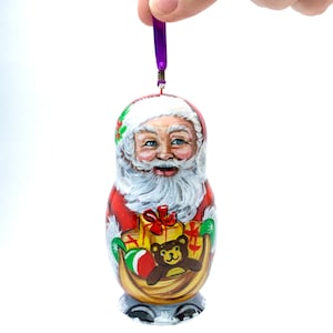 Può includere: Un ornamento natalizio in legno dipinto a mano che raffigura Babbo Natale con un abito rosso, che porta un sacco di regali e un orsacchiotto in una slitta. L'ornamento è decorato con agrifoglio verde e bacche rosse.