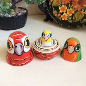 Peut inclure: Trois poup&eacute;es gigognes en bois peintes &agrave; la main, ou poup&eacute;es matriochkas, repr&eacute;sentant des oiseaux color&eacute;s. La plus grande poup&eacute;e est rouge avec des accents jaunes et orange, la poup&eacute;e du milieu est jaune avec des accents bleus et roses, et la plus petite poup&eacute;e est orange avec des accents verts.
