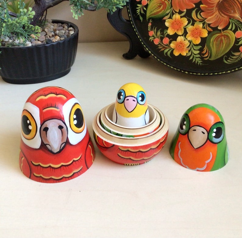 Peut inclure: Trois poup&eacute;es gigognes en bois peintes &agrave; la main, ou poup&eacute;es matriochkas, repr&eacute;sentant des oiseaux color&eacute;s. La plus grande poup&eacute;e est rouge avec des accents jaunes et orange, la poup&eacute;e du milieu est jaune avec des accents bleus et roses, et la plus petite poup&eacute;e est orange avec des accents verts.