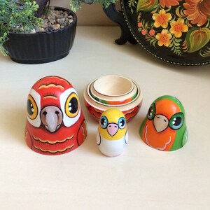 Peut inclure: Un ensemble de quatre poup&eacute;es gigognes, chacune repr&eacute;sentant un oiseau diff&eacute;rent. La plus grande poup&eacute;e est rouge avec un bec blanc et des yeux jaunes. La poup&eacute;e suivante est un oiseau jaune avec des accents roses. La troisi&egrave;me poup&eacute;e est un oiseau vert et orange avec des yeux noirs. La plus petite poup&eacute;e est une base en bois.