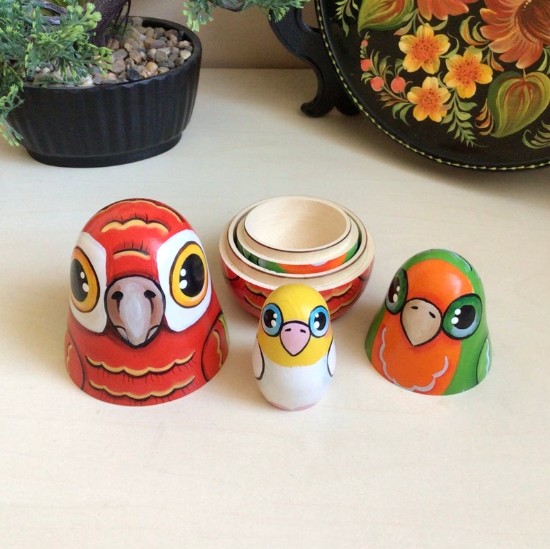 Peut inclure: Un ensemble de quatre poup&eacute;es gigognes, chacune repr&eacute;sentant un oiseau diff&eacute;rent. La plus grande poup&eacute;e est rouge avec un bec blanc et des yeux jaunes. La poup&eacute;e suivante est un oiseau jaune avec des accents roses. La troisi&egrave;me poup&eacute;e est un oiseau vert et orange avec des yeux noirs. La plus petite poup&eacute;e est une base en bois.