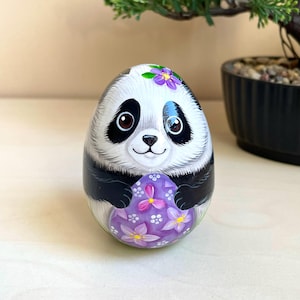 Może przedstawiać: Ręcznie malowany ceramiczny ornament w kształcie jajka przedstawiający pandę trzymającą fioletowe jajko z białymi kwiatami. Panda ma czarne oczy i białą twarz z czarnymi oznaczeniami. Ornament jest ozdobiony fioletowymi kwiatami.