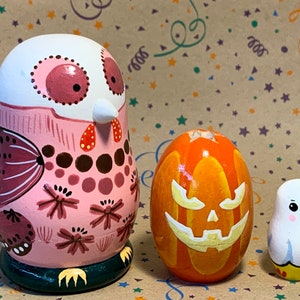 Peut inclure: Trois poupées gigognes en bois peintes à la main, ou poupées matriochka, représentant un hibou rose, une citrouille orange avec un visage de citrouille sculptée et un fantôme blanc. Les poupées sont toutes décorées de détails colorés.