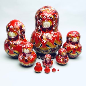 Puede incluir: Un conjunto de ocho mu&ntilde;ecas rusas tradicionales, tambi&eacute;n conocidas como mu&ntilde;ecas matrioska. Las mu&ntilde;ecas est&aacute;n pintadas de un rojo vibrante con intrincados dise&ntilde;os florales en oro y blanco. La mu&ntilde;eca m&aacute;s grande est&aacute; en la parte posterior, y la mu&ntilde;eca m&aacute;s peque&ntilde;a est&aacute; en la parte delantera.