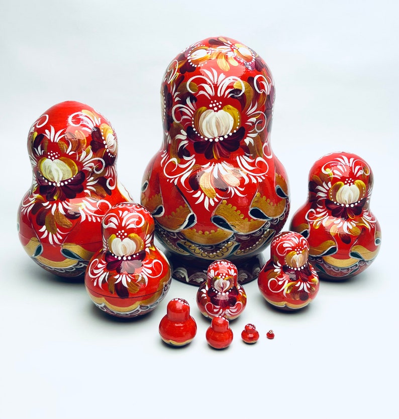 Puede incluir: Un conjunto de ocho mu&ntilde;ecas rusas tradicionales, tambi&eacute;n conocidas como mu&ntilde;ecas matrioska. Las mu&ntilde;ecas est&aacute;n pintadas de un rojo vibrante con intrincados dise&ntilde;os florales en oro y blanco. La mu&ntilde;eca m&aacute;s grande est&aacute; en la parte posterior, y la mu&ntilde;eca m&aacute;s peque&ntilde;a est&aacute; en la parte delantera.