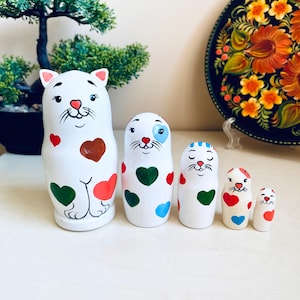 May include: A set of five white wooden nesting dolls, each painted with a different cat face and colorful hearts. The largest doll is the tallest and has a green, red, and brown heart pattern. The other dolls have green, blue, and red hearts.