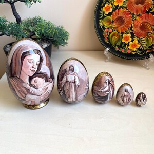 Peut inclure: Ensemble d'œufs de Pâques peints à la main, chacun avec des images religieuses. Le plus grand œuf représente une mère et un enfant, tandis que d'autres présentent des personnages en tons sépia. Les œufs varient en taille.