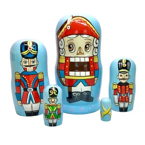 May include: A set of six hand-painted wooden nesting dolls, each depicting a nutcracker soldier in a variety of sizes. The dolls feature a light blue background with red, black, gold, and white details. The largest doll is approximately 6 inches tall.