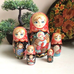 May include: A set of five traditional Russian nesting dolls, or matryoshka dolls, painted with a Christmas theme. The largest doll is red with a black background and depicts an angel holding a Christmas tree. The other dolls feature various Christmas characters, including Santa Claus, a snowman, and a reindeer.