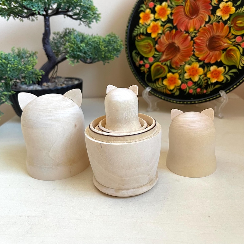 May include: Three wooden nesting dolls, also known as matryoshka dolls, with a cat-like face design. The dolls are unfinished and have a natural wood color. The largest doll is on the left, the middle doll is in the center, and the smallest doll is on the right.