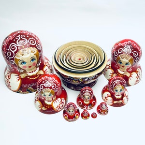 Puede incluir: Un conjunto de mu&ntilde;ecas rusas tradicionales, o mu&ntilde;ecas matrioska, con intrincados dise&ntilde;os pintados. Las mu&ntilde;ecas est&aacute;n dispuestas en c&iacute;rculo, con la mu&ntilde;eca m&aacute;s grande en el centro y la m&aacute;s peque&ntilde;a a la derecha. Las mu&ntilde;ecas est&aacute;n todas pintadas en tonos de rojo, blanco y dorado.