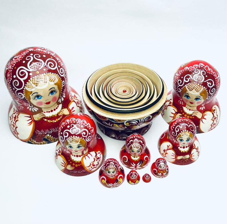 Puede incluir: Un conjunto de mu&ntilde;ecas rusas tradicionales, o mu&ntilde;ecas matrioska, con intrincados dise&ntilde;os pintados. Las mu&ntilde;ecas est&aacute;n dispuestas en c&iacute;rculo, con la mu&ntilde;eca m&aacute;s grande en el centro y la m&aacute;s peque&ntilde;a a la derecha. Las mu&ntilde;ecas est&aacute;n todas pintadas en tonos de rojo, blanco y dorado.
