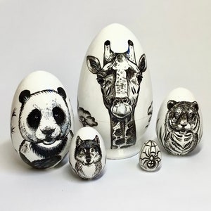 Peut inclure: Ensemble de cinq œufs en bois emboîtables peints à la main. Le plus grand œuf représente une girafe noire et blanche, le suivant une panda noire et blanche, le suivant un loup noir et blanc, le suivant un tigre noir et blanc et le plus petit œuf représente une araignée noire.