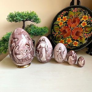 Peut inclure: Ensemble de six œufs de Pâques décoratifs avec des images religieuses. Les œufs sont peints en nuances de marron et de blanc, représentant des scènes comme la Nativité et Jésus. Un plateau rond noir avec des motifs floraux est en arrière-plan.